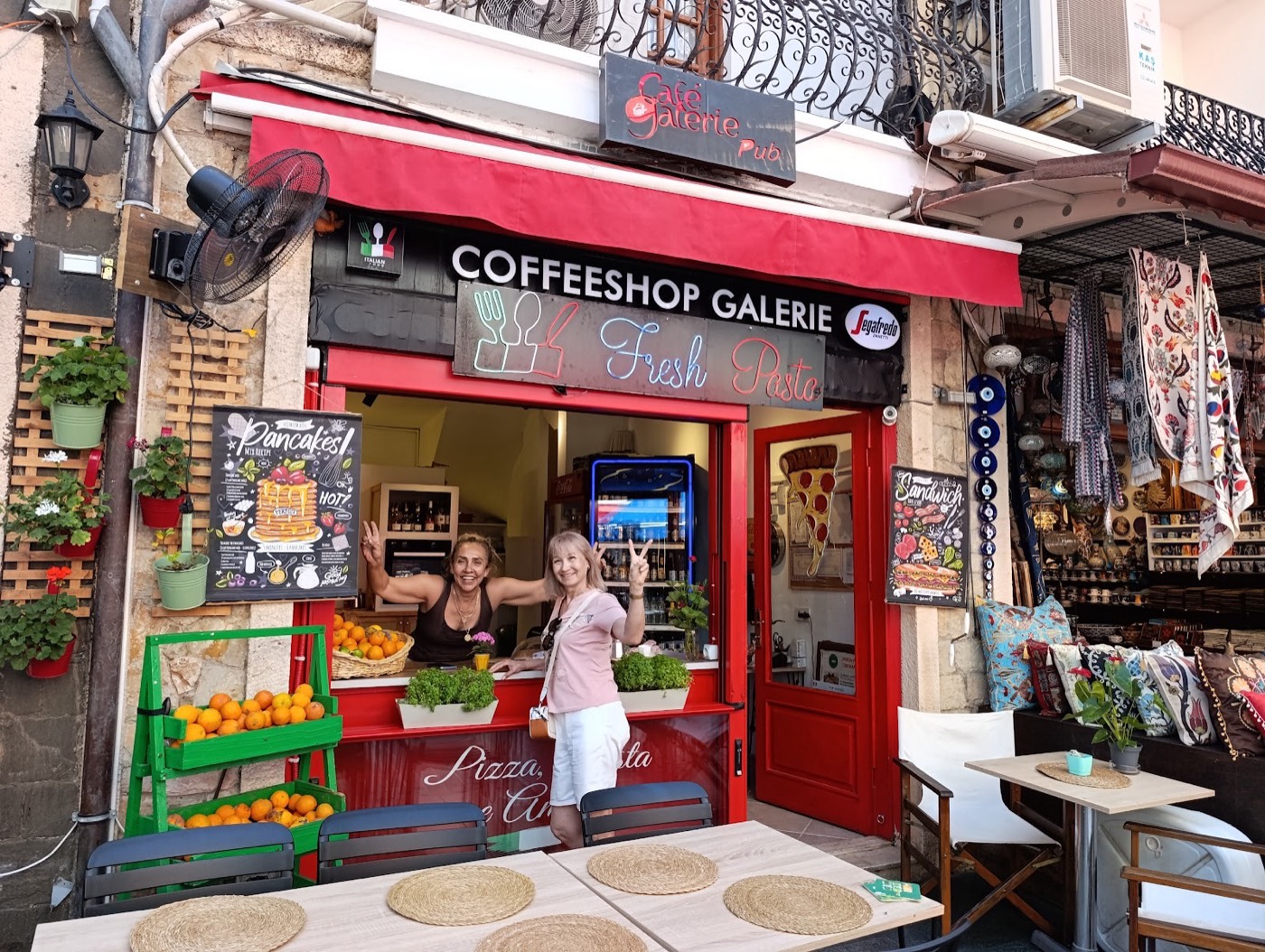 Coffee Shop Galerie'nin kırmızı tenteli cephesi, portakal sehpası ve kapıda ev sahipleri